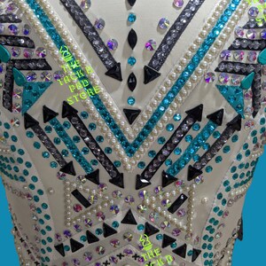 May include: White show shirt with a geometric pattern of black, teal, and silver rhinestones. The shirt is decorated with white pearls and features the text "The Tack Pad Store" on the front.