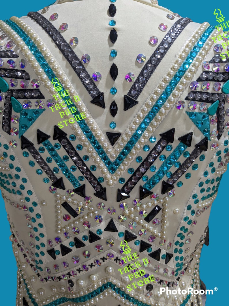 May include: White show shirt with a geometric pattern of black, teal, and silver rhinestones. The shirt is decorated with white pearls and features the text "The Tack Pad Store" on the front.