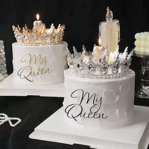 Op de afbeelding: Twee witte taarten met kroontoppers en hartvormige kaarsen. Eén kroon is goudkleurig, de andere zilverkleurig. Op de taarten staat "My Queen". Op de achtergrond is een brandende kaars te zien.