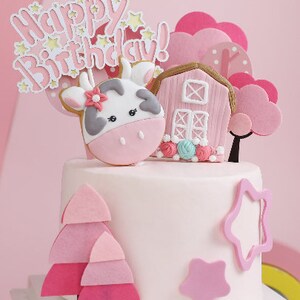 May include: A pink and white birthday cake decorated with a cow, a pink barn, pink trees, and a pink star. The cake is topped with a pink and white banner that says "Happy Birthday!"