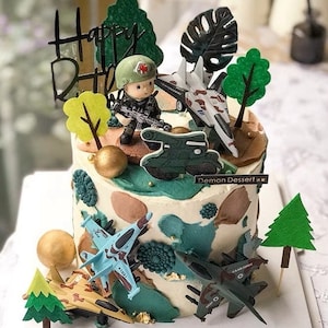 May include: A white cake decorated with green, brown, and gold accents. The cake features a miniature soldier figurine, a tank, and several toy airplanes. The cake is topped with a sign that reads "Demon Dessert".