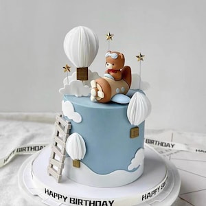 May include: A light blue birthday cake decorated with a teddy bear pilot in a biplane, white clouds, and hot air balloons. The cake has a white base with a ribbon that says "Happy Birthday". Gold stars and a ladder complete the design.