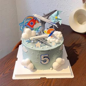 May include: A light blue birthday cake decorated with airplane and cloud toppers. The cake has the number "5" on the front and is on a white cake board. A rocket and rainbow are also on top of the cake.