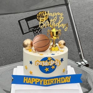 May include: A round white cake decorated with a basketball theme, featuring a gold trophy, a brown basketball, and a black basketball hoop topper. The cake includes gold and blue star accents, a blue banner with the text HAPPY BIRTHDAY, and Chinese characters.