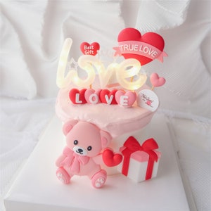 Könnte beinhalten: Ein rosa und weißer Kuchen, verziert mit einem rosa Teddybär, einer roten Geschenkbox und einem rosa und weißen 'LOVE'-Schild mit Lichtern. Der Kuchen hat auch ein rotes Herz mit dem Text 'TRUE LOVE' und ein kleines rosa Herz mit dem Text 'Best Gift'.