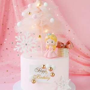 Puede incluir: Un pastel rosa y blanco decorado con un árbol de tul, un copo de nieve, una pequeña muñeca y una caja de regalo. El pastel está cubierto con glaseado blanco y tiene las palabras "Happy Birthday" escritas en él.