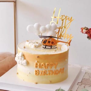 May include: A round cake with a gradient orange and yellow frosting, decorated with a toy helicopter, cloud-shaped meringues, and a gold "Happy Birthday" topper. The words "Happy Birthday" are written on the cake.