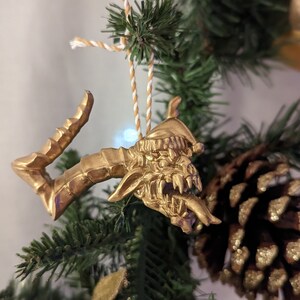 May include: A gold-coloured dragon ornament with a fierce expression, hanging from a white and gold twine on a green Christmas tree.