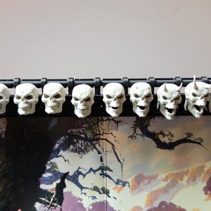 May include: A black plastic display piece featuring eleven white skull figurines. The skulls are arranged in a row, with some having horns. The skulls are detailed with eye sockets and teeth. The display piece is likely for a tabletop game.