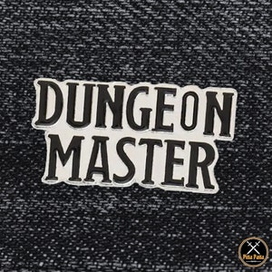 May include: A silver-coloured pin featuring the text "DUNGEON MASTER" in bold, black lettering. The rectangular pin has a black border and is set against a textured, dark grey background. A gaming accessory.