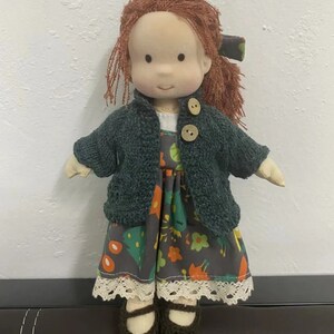 May include: A handmade doll with red yarn hair, a green knitted cardigan, a floral print dress, and brown crocheted shoes.