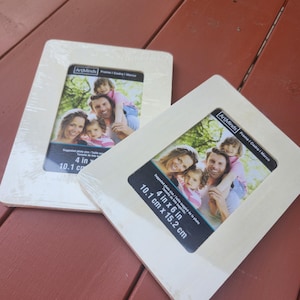 May include: Two light-colored wooden picture frames, each holding a family photo. The frames are rectangular with a square opening for a 4 in x 6 in (10.1 cm x 15.2 cm) photo. The frames are packaged in clear plastic.