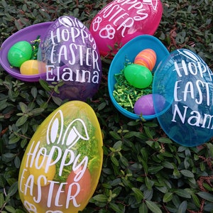 May include: Four clear plastic Easter eggs with bunny ears and the words "Hoppy Easter" on them. The eggs are in different colors: pink, purple, blue, and yellow. Each egg has a different name on it: "Hoppy Easter", "Hoppy Easter Elanna", "Hoppy Easter Nam", and "Hoppy Easter".