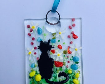 May include: A glass ornament featuring a black cat silhouette sitting in a field of white flowers with red, blue, and green accents.