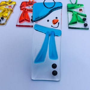 May include: Rectangular glass snowman decorations with blue, orange, and black details. Each decoration has a different coloured scarf and hat. The decorations are strung with colourful ribbons for hanging. The snowman's face is made with black dots for eyes and a curved line for a smile.