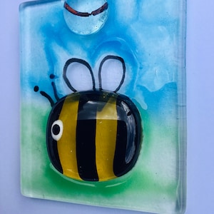 May include: A glass suncatcher featuring a cartoon bee with black and yellow stripes. The bee is on a green and blue background with a clear glass bead hanging from the top.