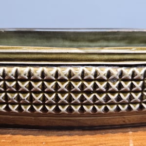 Puede incluir: Un plato rectangular de cerámica verde oliva con un patrón geométrico texturizado en los lados. El plato tiene un acabado brillante y un interior liso y oscuro. El diseño es un patrón repetitivo de formas de diamante en relieve.