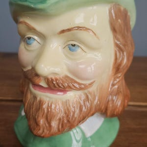 Może przedstawiać: Ceramiczny kubek w kształcie głowy mężczyzny z zielonym kapeluszem, brązową brodą i niebieskimi oczami. Kubek jest pomalowany realistycznymi detalami i ma fantazyjny design.