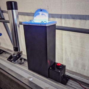 May include: A black box with a blue light inside and a red button on the side. The box is mounted on a metal surface.