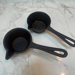 Pode incluir: Duas pequenas panelas de molho de ferro fundido preto com cabos longos e bicos. As panelas são redondas com uma pequena borda para despejar. As alças têm um orifício na extremidade para pendurar. As panelas estão em uma superfície de mármore.