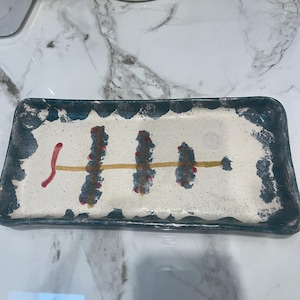 May include: Rectangular ceramic serving tray with a white base and a dark teal border. The tray features a yellow horizontal line with red and teal vertical accents. The tray is likely handmade and has a rustic aesthetic.