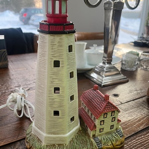 May include: A ceramic lighthouse lamp with a red top and a small attached building. The lighthouse is cream-colored with black accents and square windows. The base is green and brown, resembling a grassy area.