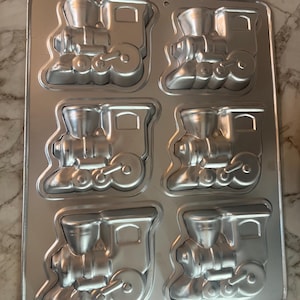 Könnte beinhalten: Eine rechteckige, silberfarbene Backform aus Metall mit sechs zugförmigen Formen. Jede Form weist ein detailliertes Lokomotivdesign auf, einschließlich Schornstein, Führerhaus und Rädern. Die Form ist zum Backen von Kuchen oder anderen Leckereien bestimmt.