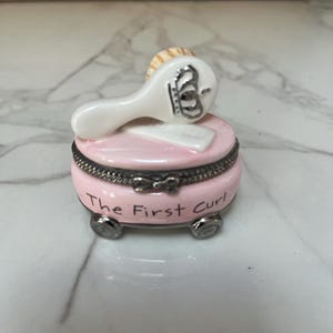 May include: A small, pink, round trinket box with silver trim and feet. The box has the words "The First Curl" in black script. A small, white brush with a silver crown design rests on top.