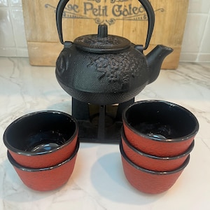 May include: A black cast iron teapot with a matching stand and two stacks of red and black tea cups. The teapot has a floral design and a curved handle. The tea cups are red on the outside and black on the inside.