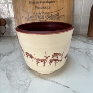 May include: Cream-colored ceramic bowl with a dark red rim and deer illustrations. The bowl has a wide opening and a slightly flared shape. The deer are depicted in a simple, brown line drawing style. The background includes a wooden sign.