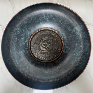 May include: A large, round, dark teal metal bowl with a raised rim and a central medallion. The medallion features a detailed relief of a seated figure and text. The bowl has a textured, mottled appearance with copper-colored accents.