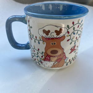 May include: Ceramic mug with a blue handle and rim, featuring a cartoon reindeer chef design. The reindeer wears a chef's hat and scarf, holding a spoon. The mug is decorated with colorful Christmas lights.