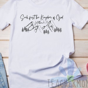 May include: White t-shirt with a black graphic of mountains and trees. The text "Seek first the Kingdom of God" and "Matthew 6:33" is printed on the shirt.