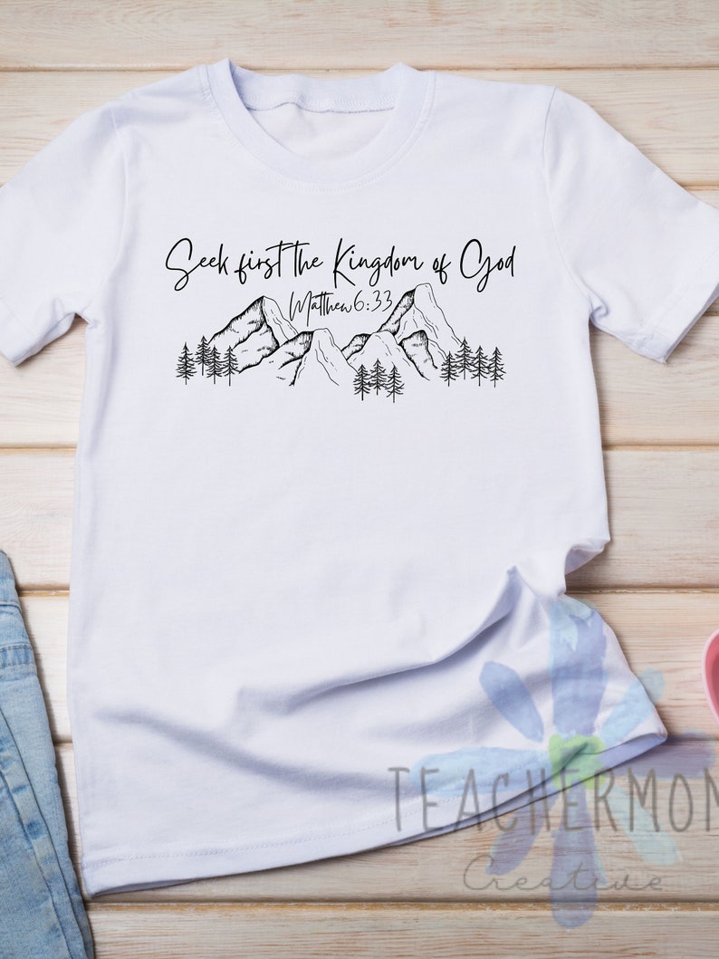 May include: White t-shirt with a black graphic of mountains and trees. The text "Seek first the Kingdom of God" and "Matthew 6:33" is printed on the shirt.