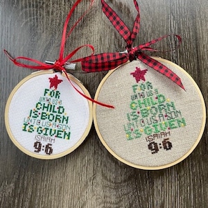 Puede incluir: Dos adornos navideños bordados con un diseño de árbol de Navidad verde y el texto "For unto us a child is born unto us a son is given Isaiah 9:6". Los adornos están sobre una superficie de madera con cinta a cuadros rojos y negros.