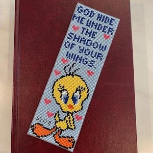 May include: A handmade bookmark featuring Tweety Bird with the text "GOD HIDE ME UNDER THE SHADOW OF YOUR WINGS." The bookmark is light blue with a cross-stitch design, and is resting on a burgundy book with a red ribbon.