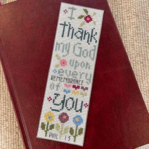 May include: A handmade cross-stitch bookmark with the text "I Thank my God upon every remembrance of you" and floral accents. The bookmark is resting on a burgundy leather-bound book with a red ribbon.
