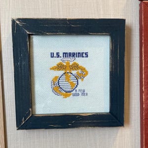 May include: Cross-stitch art in a distressed navy blue frame. The artwork displays "U.S. MARINES" above an eagle, globe, and anchor emblem, with "A FEW GOOD MEN" below. The background is a light blue fabric.