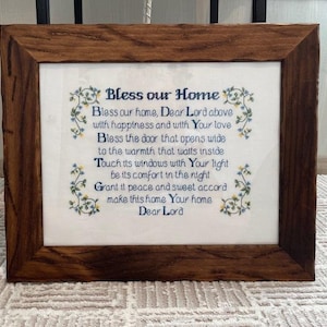 Op de afbeelding: Een geborduurde borduurwerk met een donkerbruine houten lijst. Het borduurwerk bevat de tekst "Bless our Home" in blauw schrift, samen met een gebed. Bloemenaccenten in blauw en geel versieren de randen van de tekst.