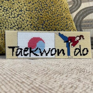 Puede incluir: Un letrero rectangular bordado a punto de cruz con la palabra "Taekwondo" en letras negras. El letrero presenta una imagen estilizada de una persona realizando una patada y la bandera coreana. El fondo es de color beige claro.