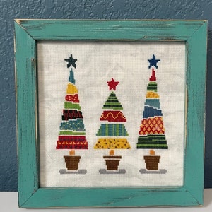 Puede incluir: Tres árboles de Navidad geométricos y coloridos con estrellas en la parte superior, enmarcados en un marco de madera turquesa.