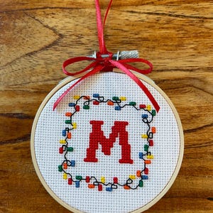 May include: A white embroidery hoop with a red ribbon attached. The hoop contains a cross-stitch design of the letter "M" surrounded by a wreath of colourful lights.