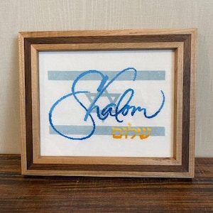 May include: A framed cross-stitch design featuring the word "Shalom" in blue cursive script with a Star of David and the Hebrew word for peace in yellow. The design is on a white background with blue and white stripes.