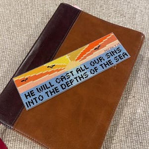 Könnte beinhalten: Ein handgefertigter Lesezeichen mit Kreuzstich-Design, das eine Sonnenuntergangsszene in Orange, Gelb und Blau zeigt. Der Text lautet: „HE WILL CAST ALL OUR SINS INTO THE DEPTHS OF THE SEA“. Das Lesezeichen liegt auf einer braunen Lederbibel.
