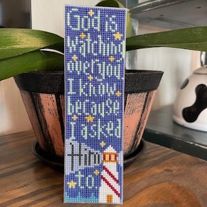 Könnte beinhalten: Handgefertigter, blauer Kreuzstich-Lesezeichen mit dem Text „God is watching over you I know because I asked Him to“. Das Lesezeichen zeigt unten ein Leuchtturm-Design und gelbe Sternakzente.