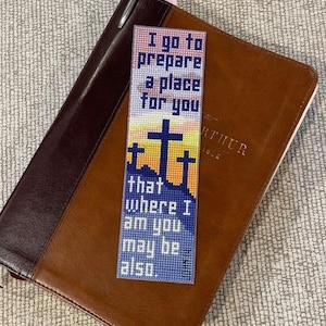 Puede incluir: Marcapáginas hecho a mano con diseño de punto de cruz. El marcapáginas presenta el texto "Voy a preparar un lugar para ti..." y tres cruces sobre un fondo de puesta de sol. El marcapáginas está sobre una Biblia de cuero marrón.