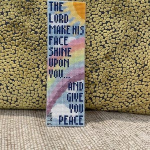 Könnte beinhalten: Ein rechteckiger, handgefertigter Lesezeichen mit dem Text "THE LORD MAKE HIS FACE SHINE UPON YOU... AND GIVE YOU PEACE." Das Lesezeichen hat ein farbenfrohes Kreuzstichdesign mit blauen, orangen, rosa und violetten Farbtönen.