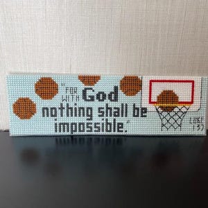 Op de afbeelding: Een kruissteekbord met een lichtblauwe achtergrond met de tekst "Want bij God is niets onmogelijk" en een basketbalring. Bruine basketballen zijn ook afgebeeld. De tekst "Lucas 1:37" is ook opgenomen.