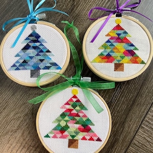 以下が含まれることがあります： カラフルな幾何学模様の木が描かれた、手作りクリスマスオーナメント3点セット。各オーナメントは白い布地に刺繍され、木製の輪で囲まれています。