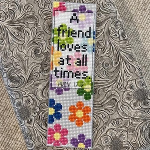 May include: A handmade bookmark with the text "A friend loves at all times" and "PROV 17:17" in black. The bookmark features colorful cross-stitched flowers in pink, orange, blue, and purple. The background is a light gray.